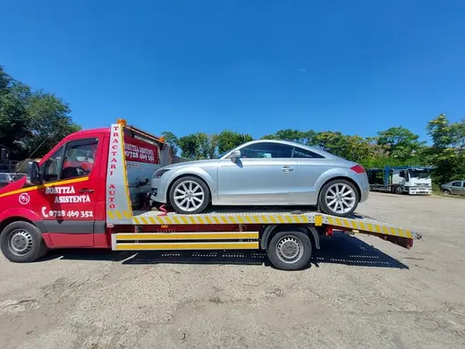 Coupe argintiu încărcat pe platformă, tractat cu utilitară roșie pe teren viran