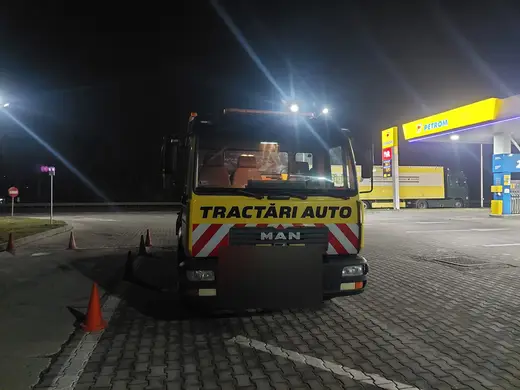 Autoutilitară galbenă de tractări, cu platformă goală, staționată noaptea lângă o benzinărie