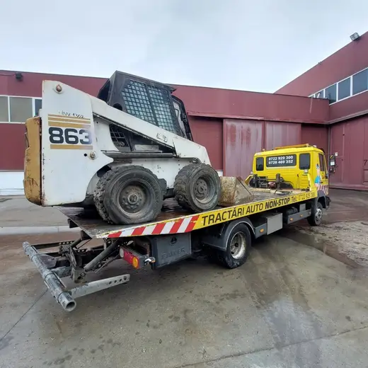 Buldoexcavator compact alb încărcat pe platforma hidraulică, lângă o hală industrială